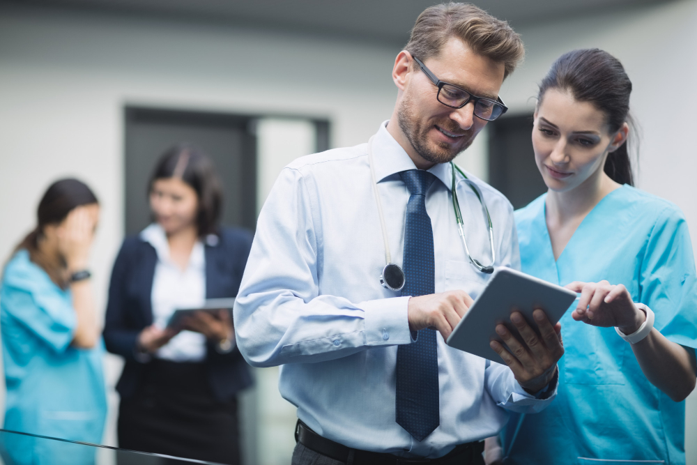 stock photo of a man with a tie holding and using a digital tablet with a woman in hospital scrubs, and a woman in the background using a tablet with another woman in hospital scrubs