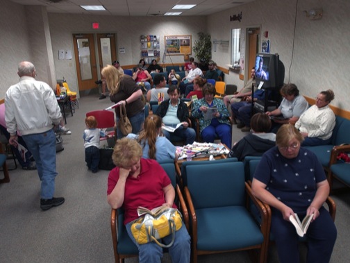 a photo of a cavernous and doctor's office with patients sitting crowded, and a mother managing a small child